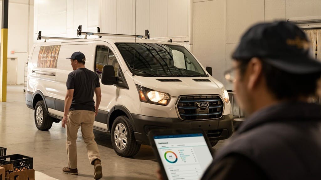 A worker walks by a Ford E-Transit while another worker in the foreground looks at data on their tablet.