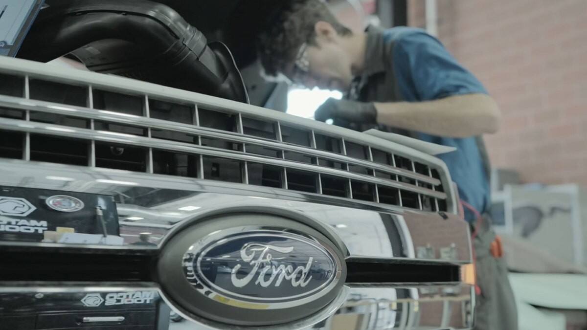 A close-up on a grill with the Ford logo as a technician works on the engine in the background